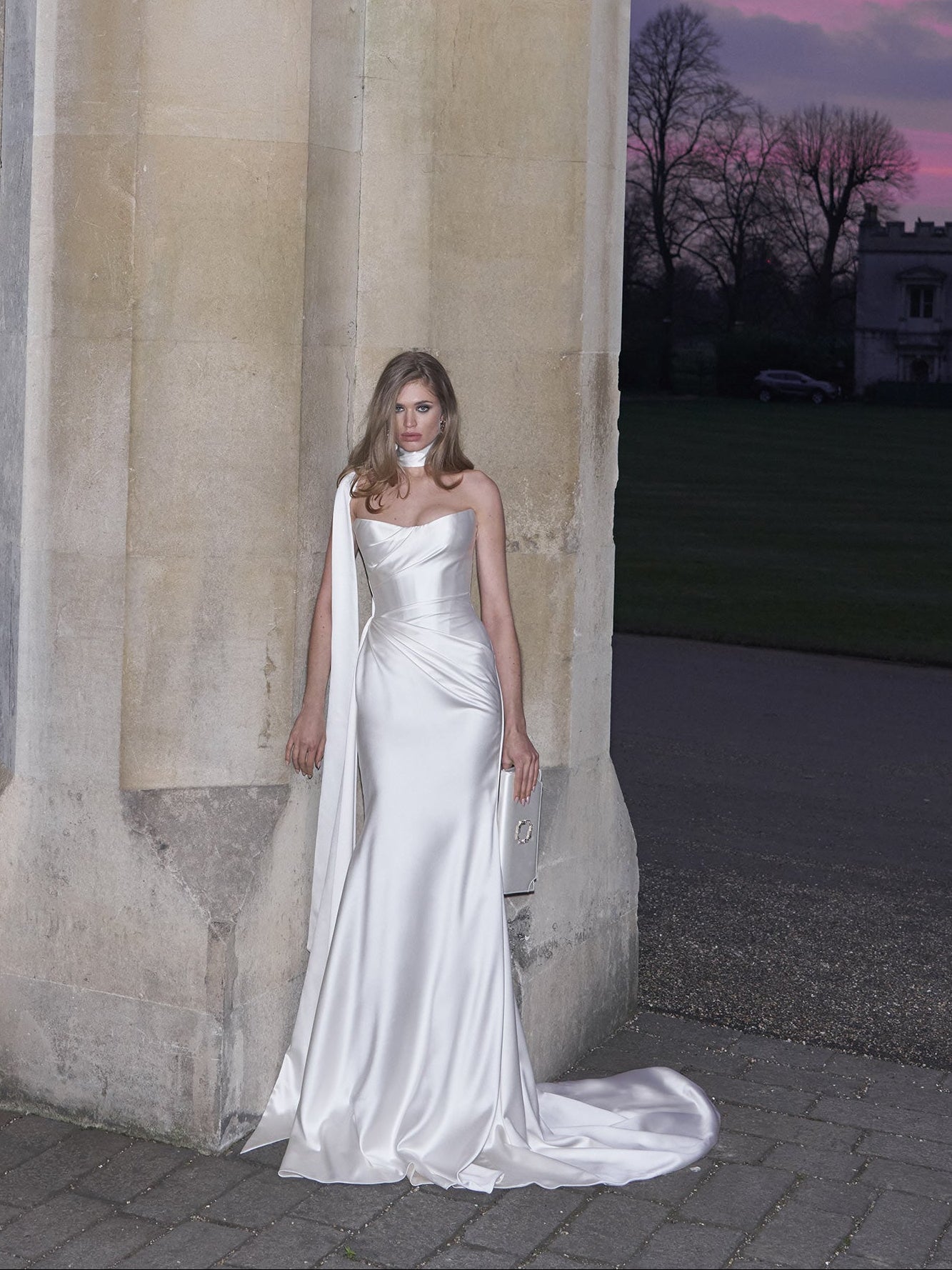 Woman in a white wedding dress standing next to stone columns with a sunset sky.