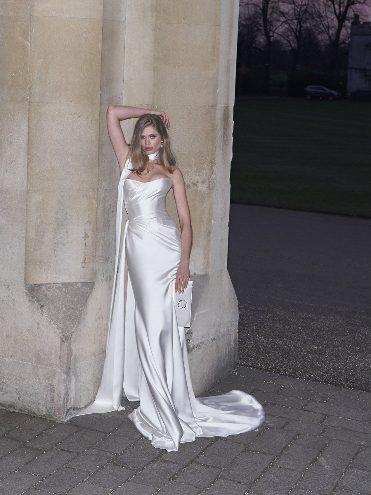 Woman in a white wedding dress standing against a stone wall with a sunset in the background.