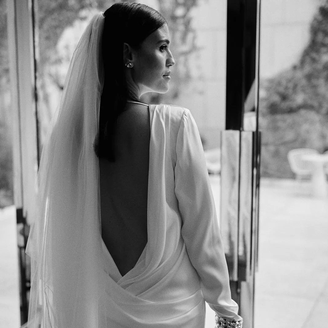 A black and white photograph of a bride from behind, wearing a long-sleeved open-back wedding gown and veil, holding a bouquet as she stands near a glass door.