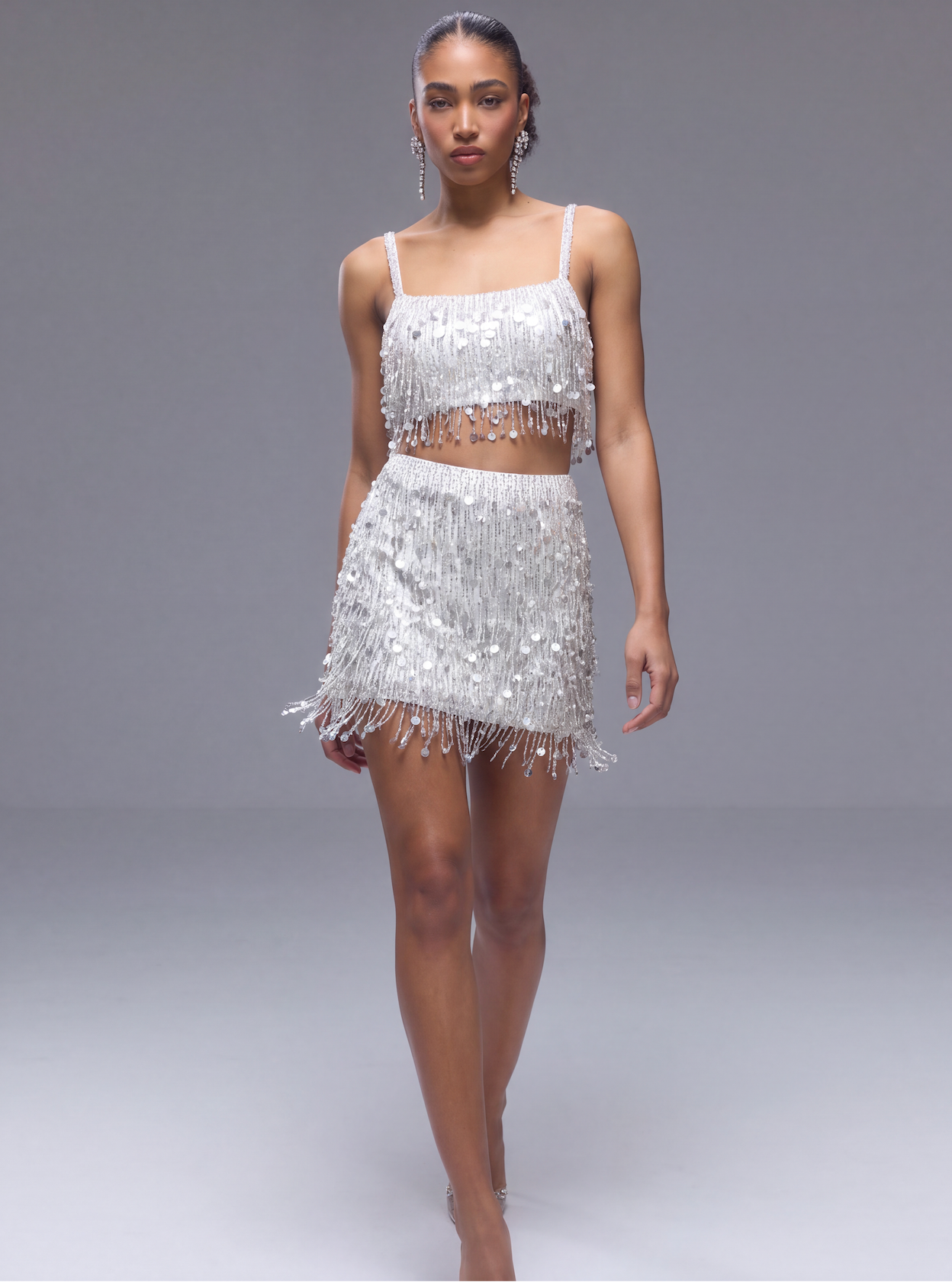 A model wearing a sparkling silver sequined two-piece outfit consisting of a fringed crop top and a matching fringed mini skirt, set against a plain gray studio background.