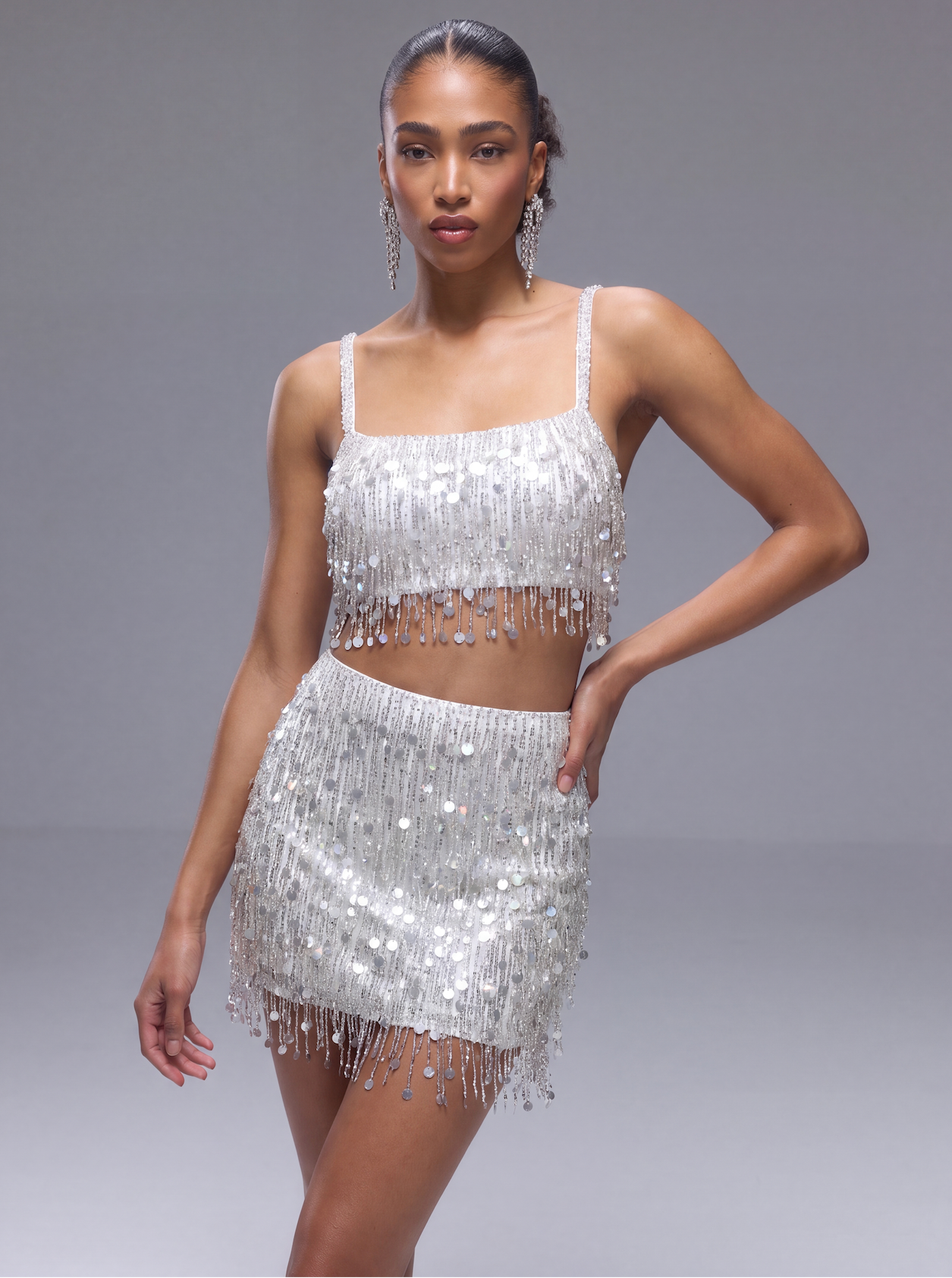 A model wearing a sparkling silver sequined two-piece set consisting of a fringed crop top and a matching fringed mini skirt, posed against a neutral gray studio background.