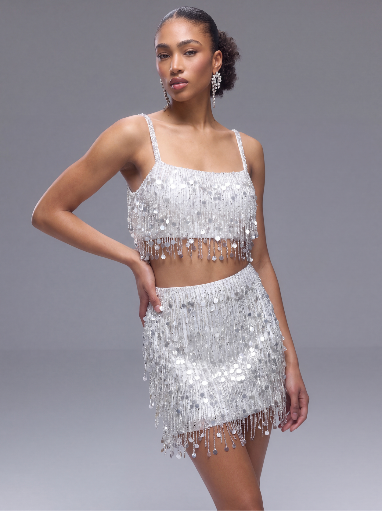 A model wears a sparkling silver sequin two-piece outfit consisting of a fringed crop top and matching mini skirt, set against a solid gray studio background.
