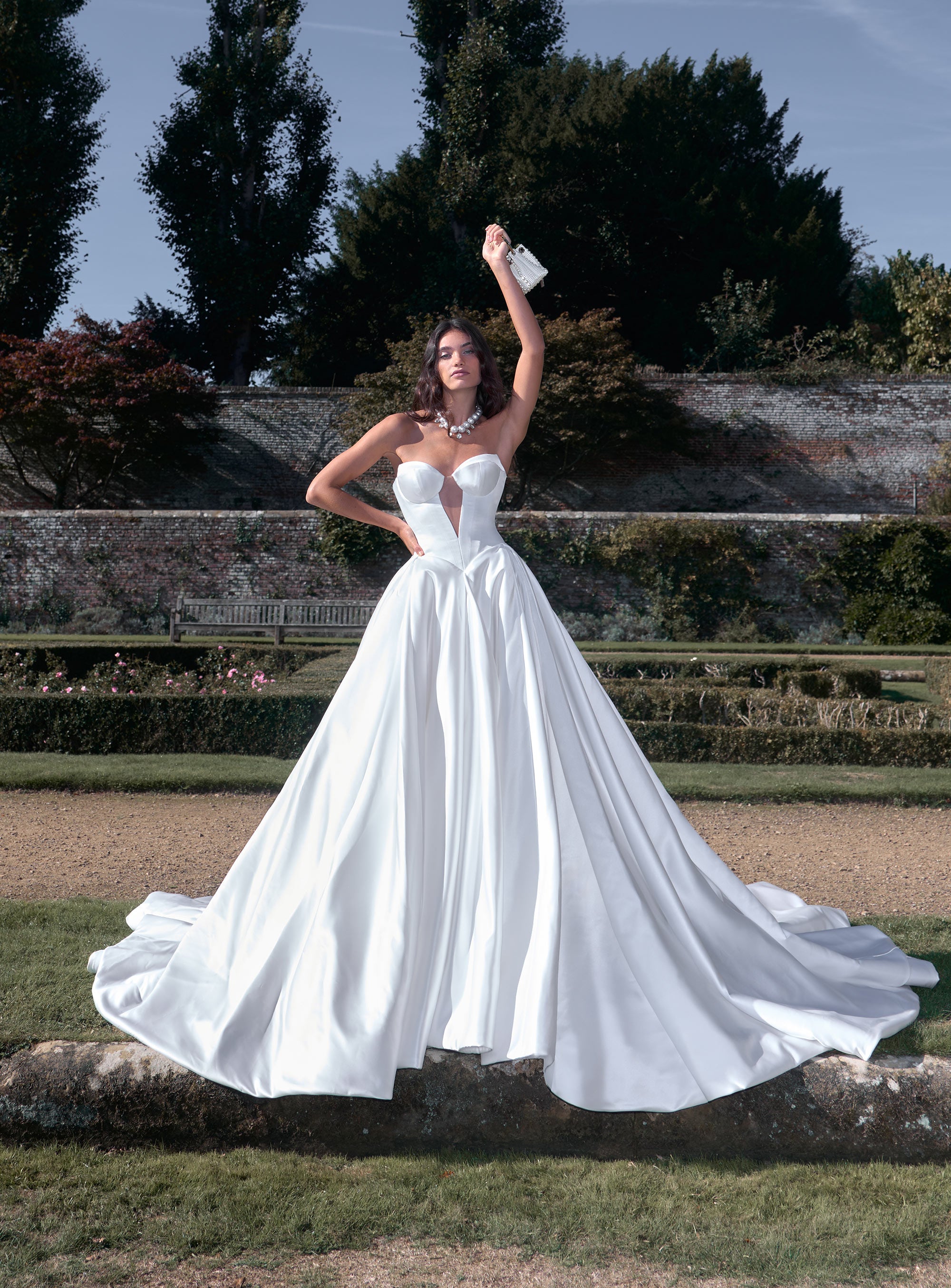 Woman in a white wedding dress standing outdoors with trees and a stone wall in the background