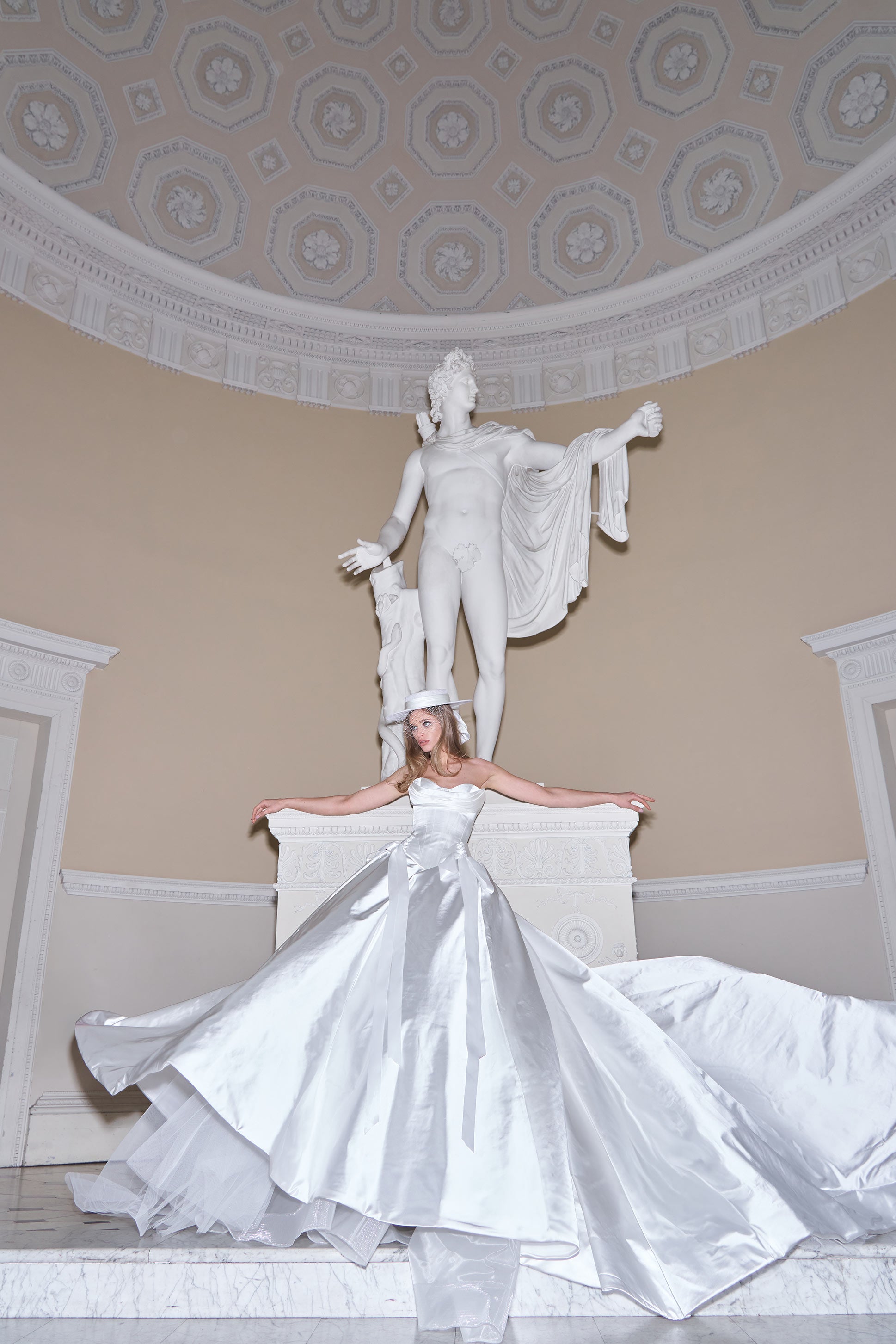Woman in a white dress standing next to a classical statue in an elegant interior setting