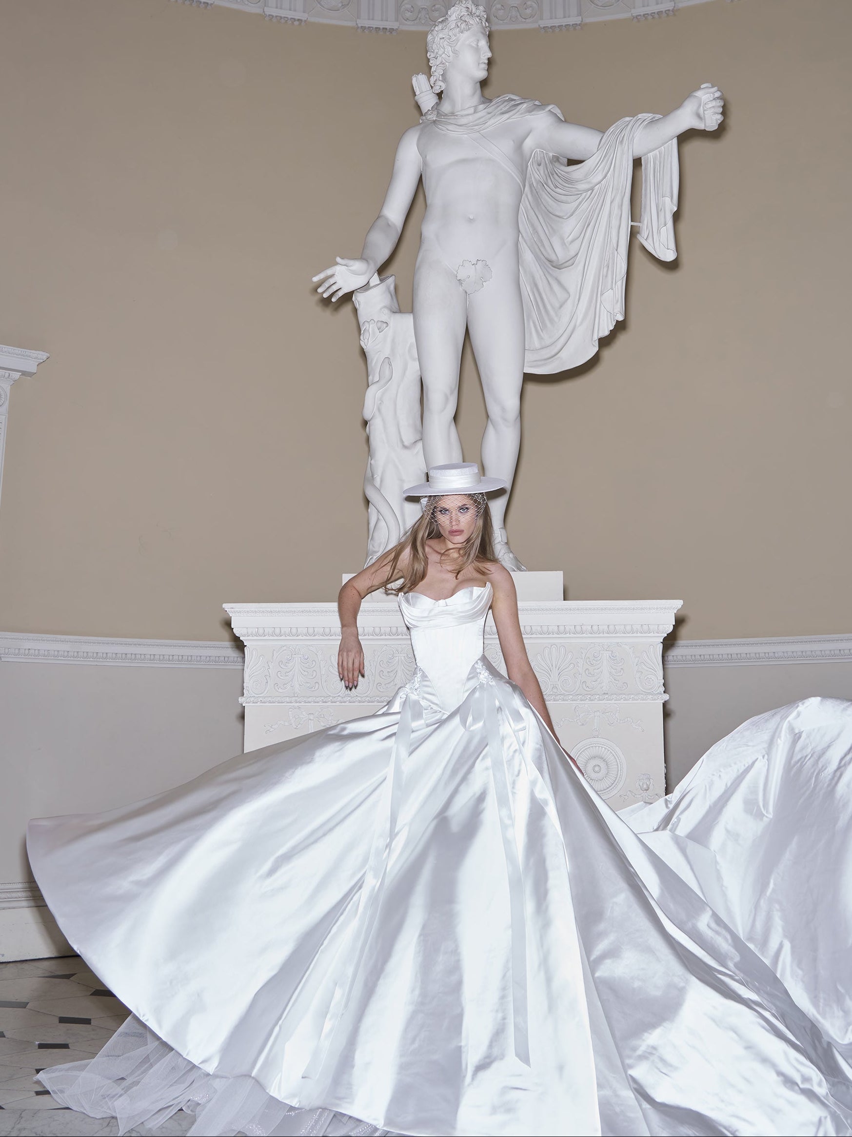 Woman in a white wedding dress standing in front of a classical statue in an elegant room.