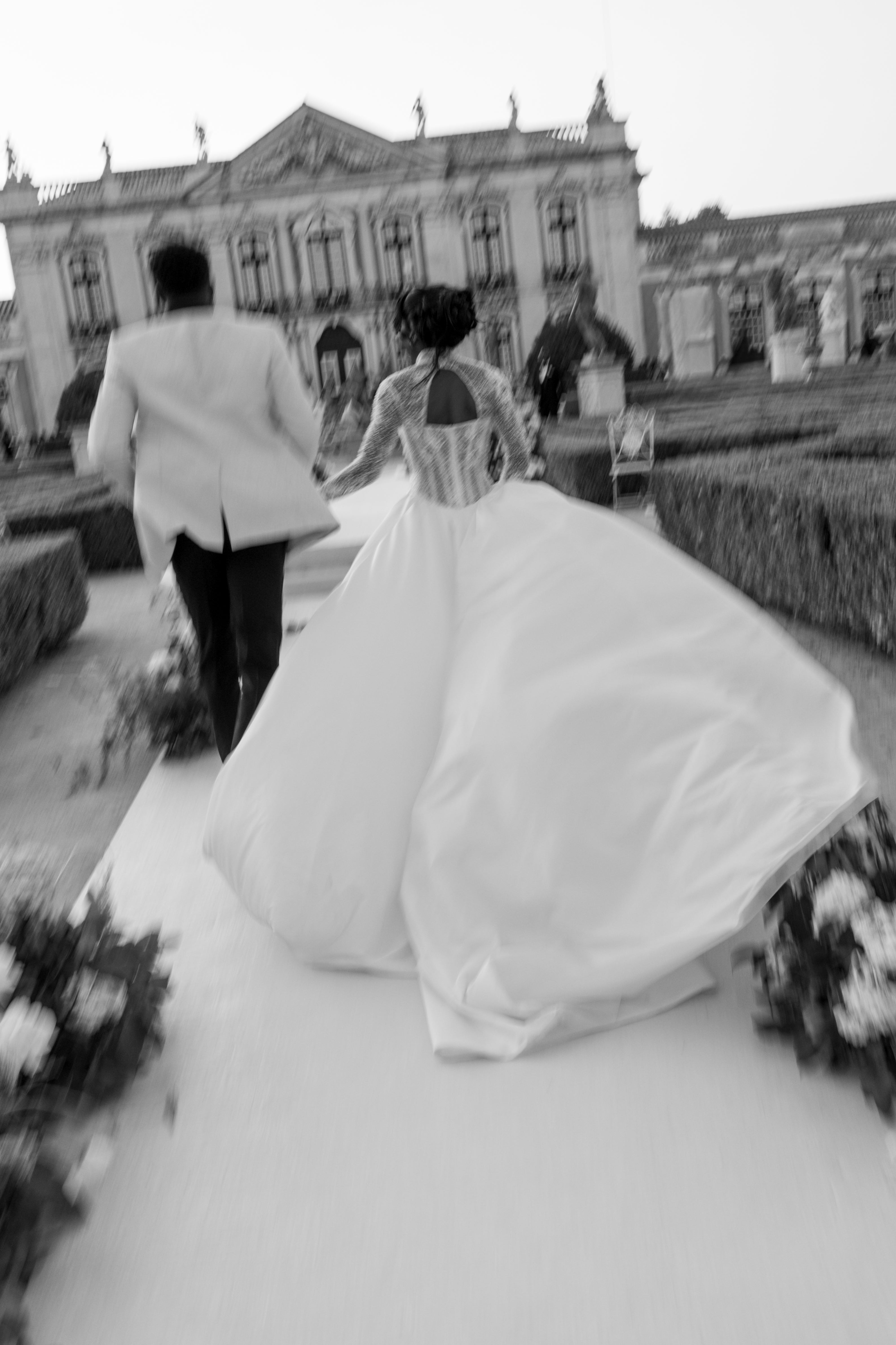 Black and white photo of a couple in wedding attire walking away from a historic building.