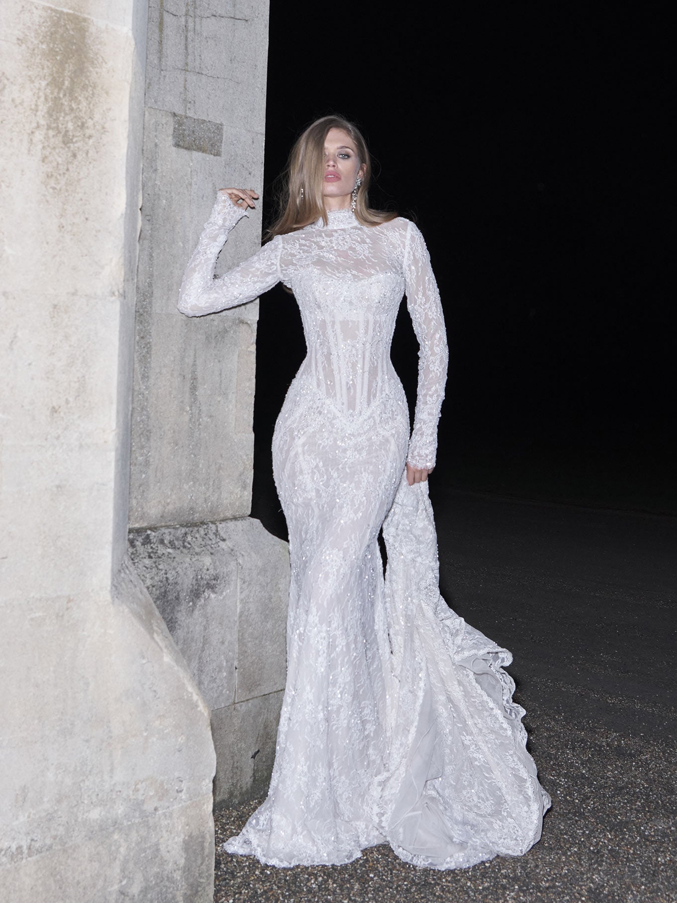 Woman in a white lace evening gown standing against a dark background.