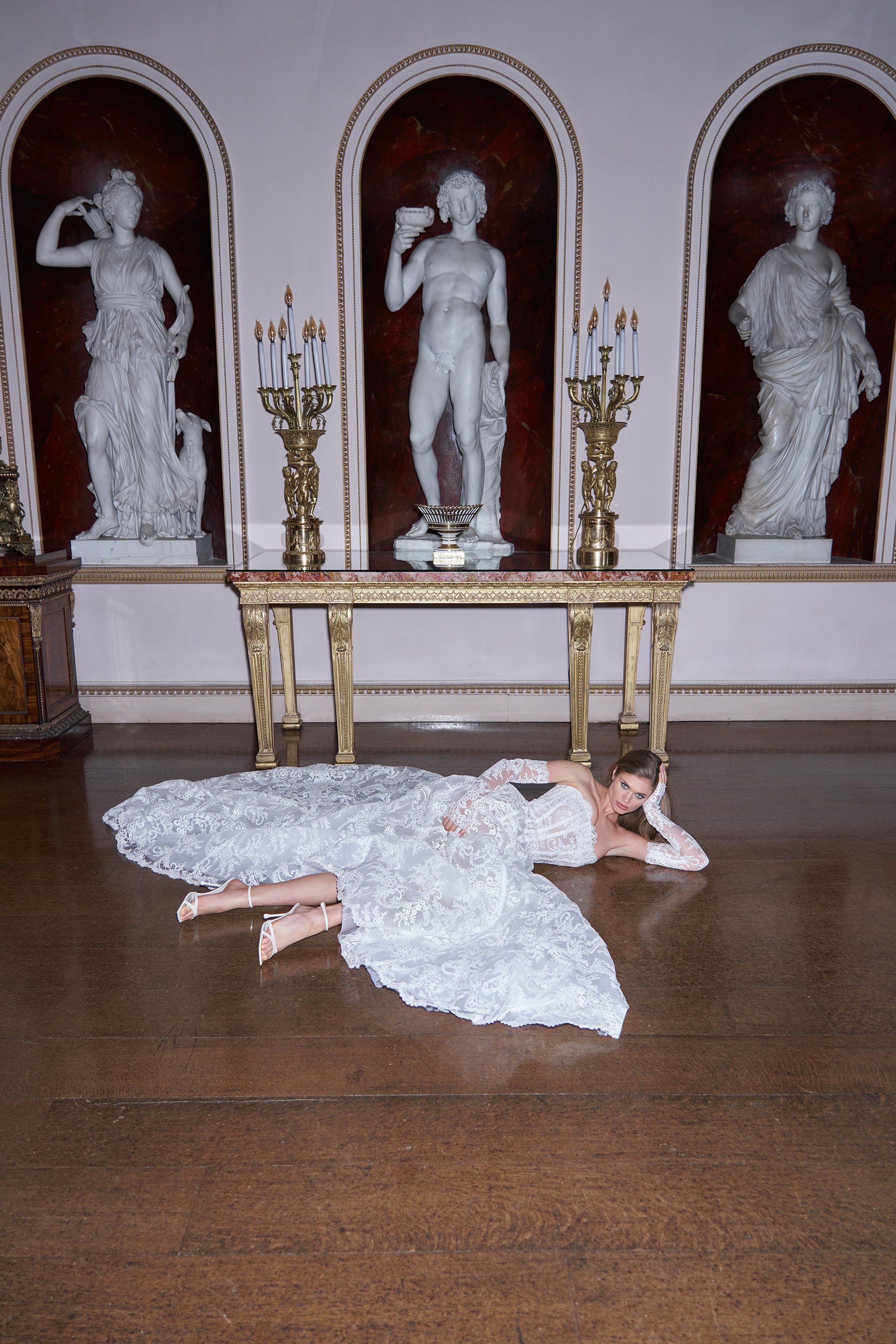 Woman in a white dress lying on the floor in front of classical statues and candelabras.