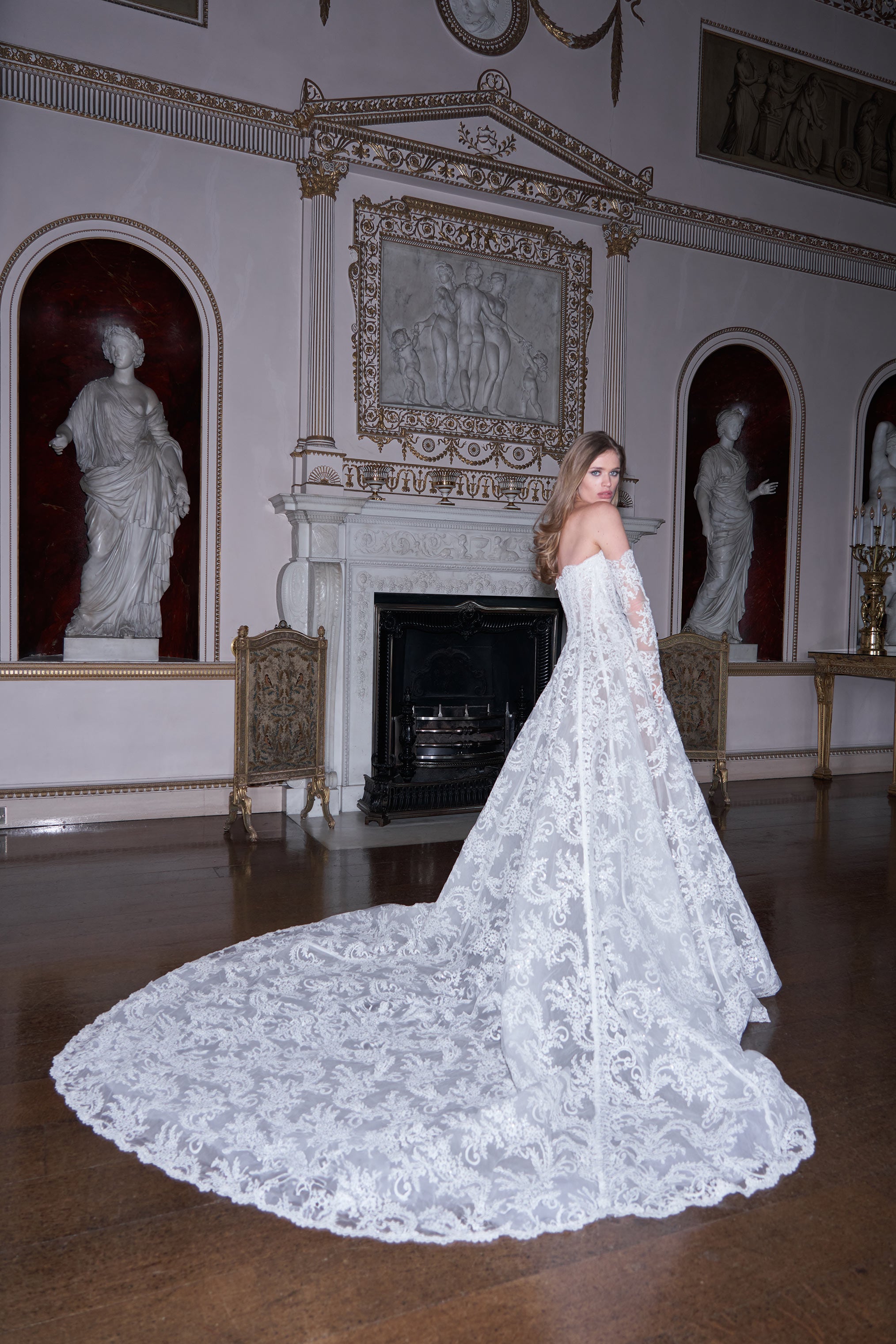 Woman in a white lace wedding dress standing in an elegant room with statues and a fireplace.