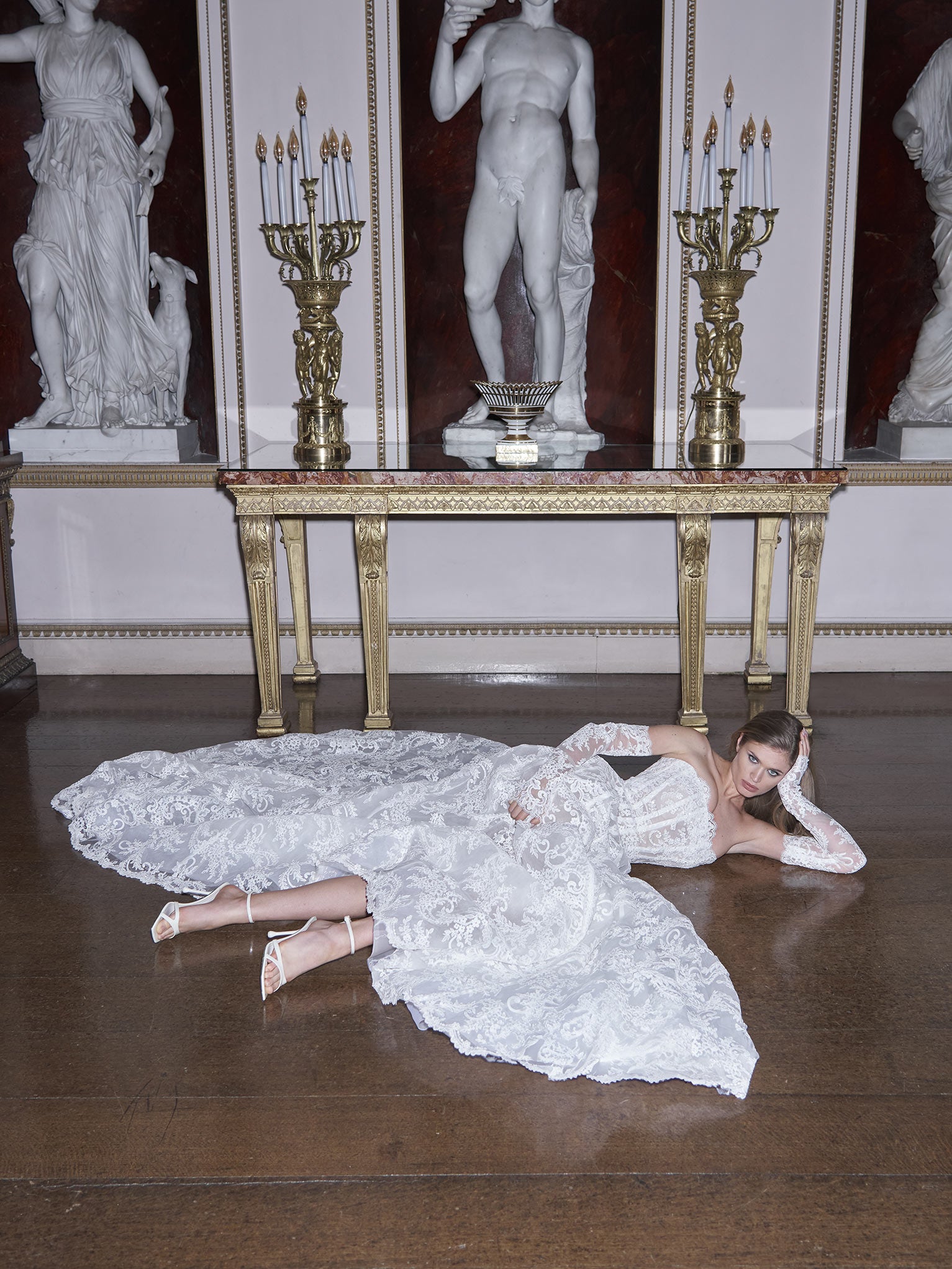 Woman in a white lace dress lying on the floor with classical statues and candelabras in the background.
