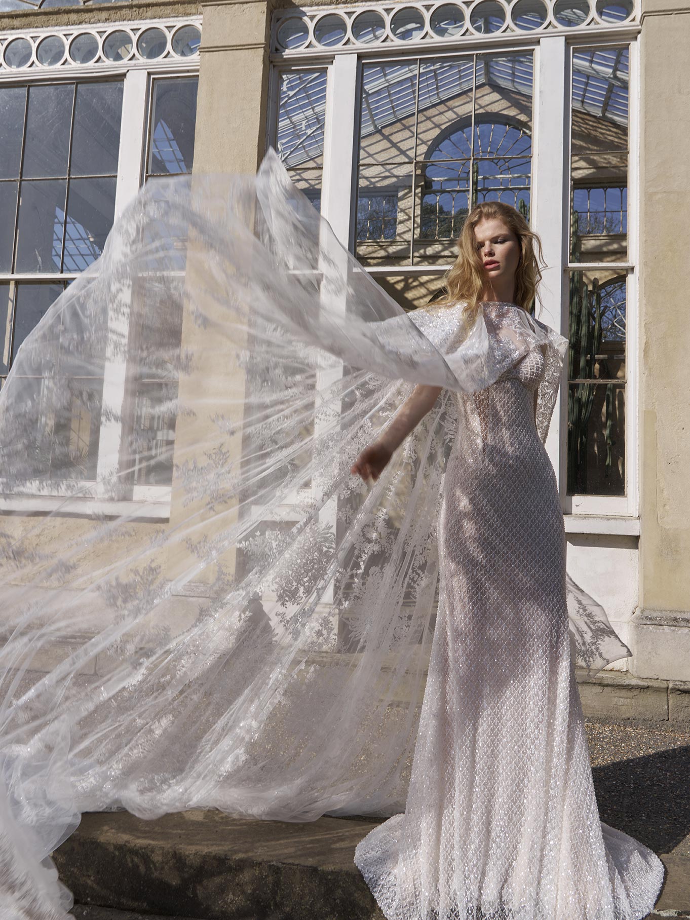 Woman in a white wedding dress with a long train standing in front of a large glass window.