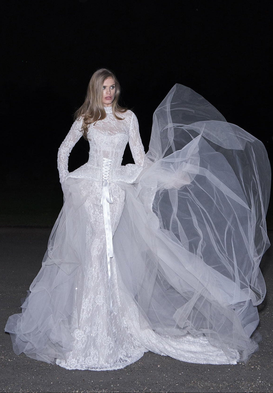 A woman wearing a long-sleeved white lace wedding gown with a sheer tulle overskirt and a long flowing veil, photographed against a dark night background.