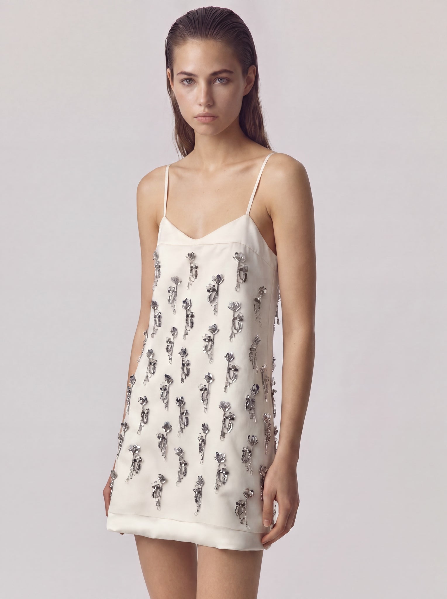 A woman modeling a cream-colored spaghetti-strap mini dress with an all-over black floral print, set against a plain light gray studio background.