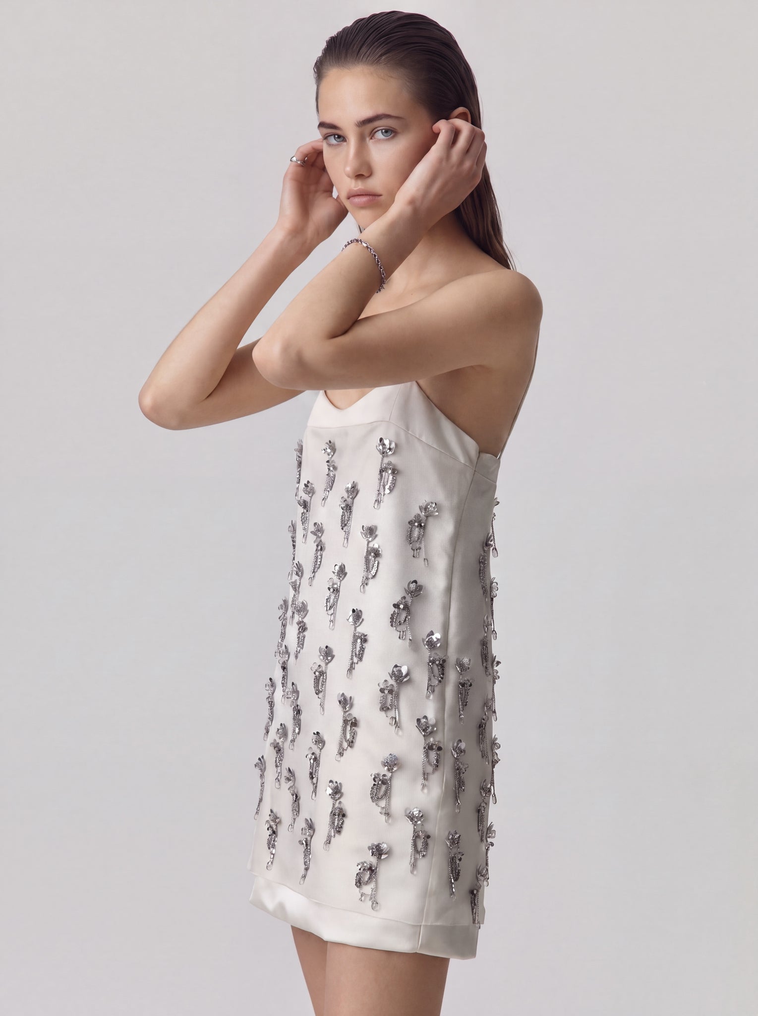 A model wearing a white sleeveless mini dress decorated with silver metallic rose-shaped embellishments, posed against a light gray studio background.