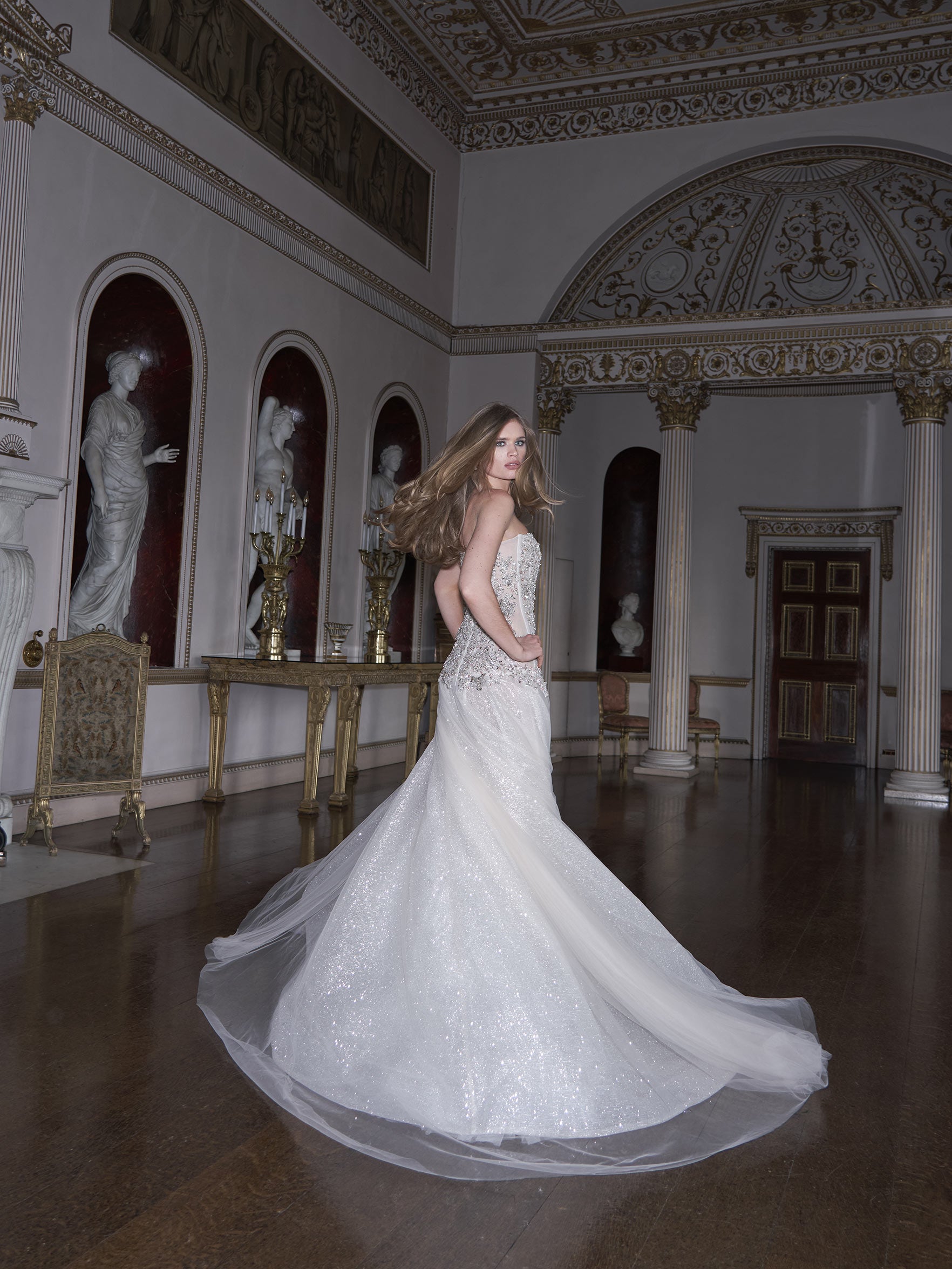 Woman in a white wedding dress standing in an ornate room with classical sculptures.