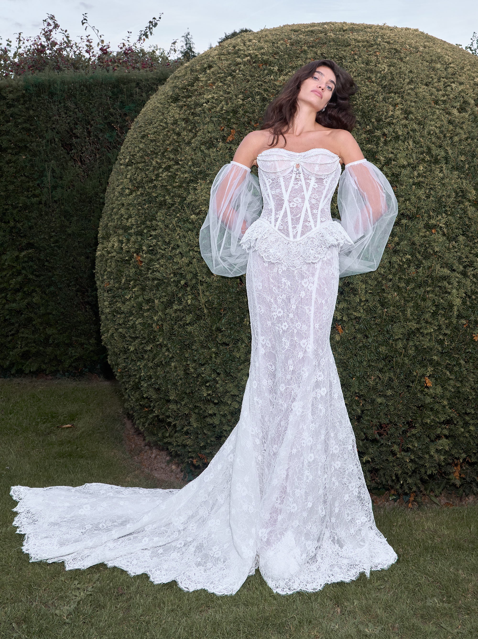 Woman in a white lace wedding dress with sheer sleeves standing in front of green bushes.