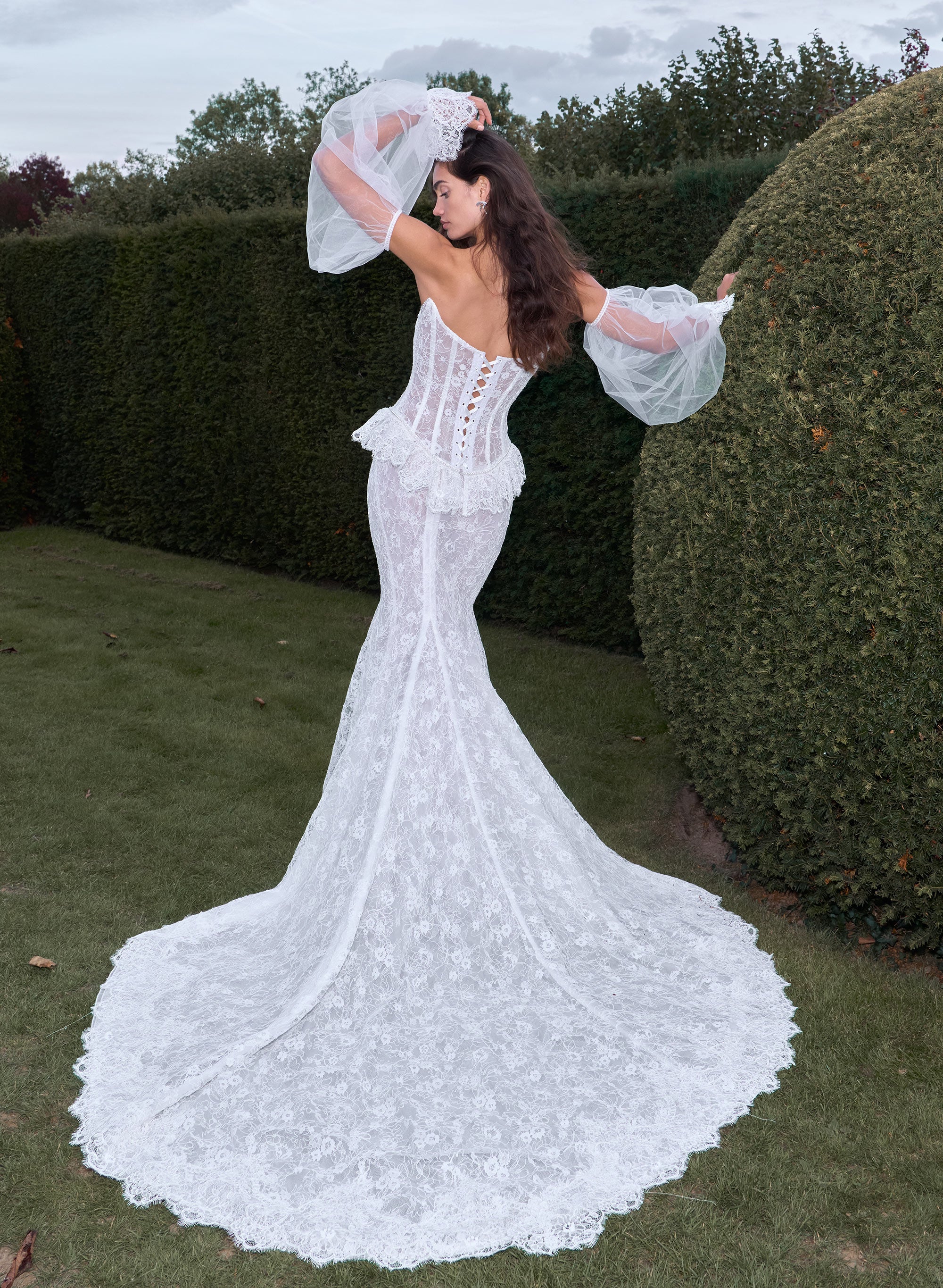 Woman in a white lace wedding dress standing outdoors with greenery in the background