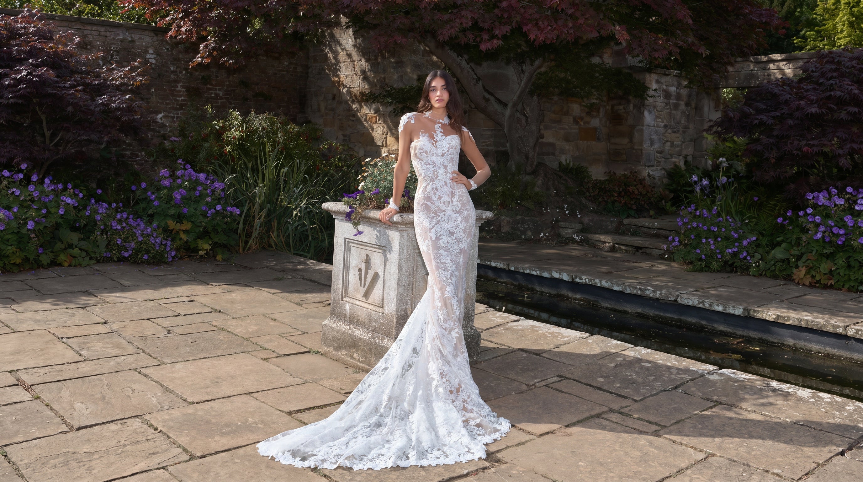 Woman in a white wedding dress standing in a garden with flowers and a stone wall in the background.