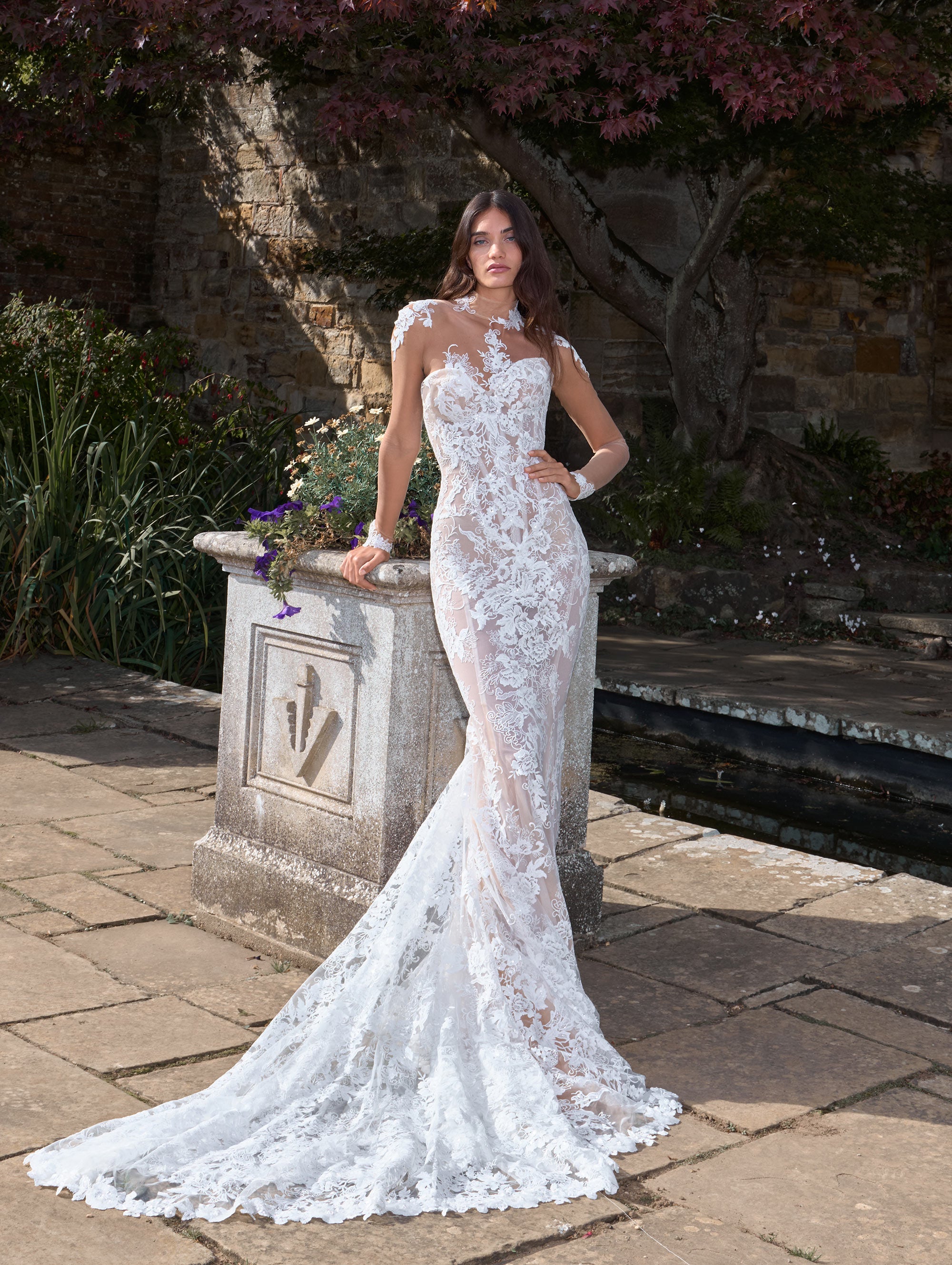 Woman in a white lace wedding dress standing outdoors near a stone structure.