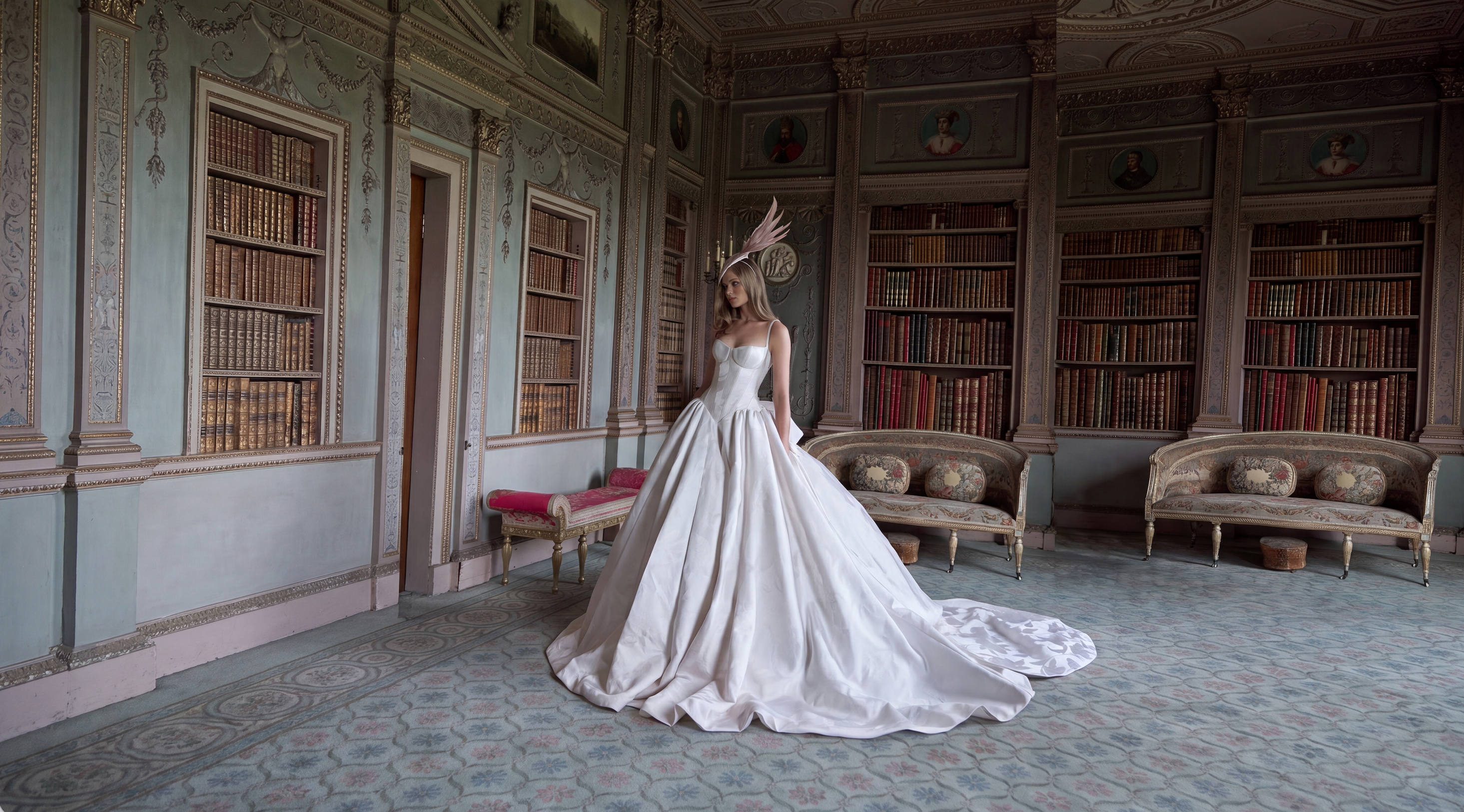 A woman wearing a voluminous white bridal gown with a dramatic feathered headpiece stands in a grand, old book-filled room with ornate furniture.
