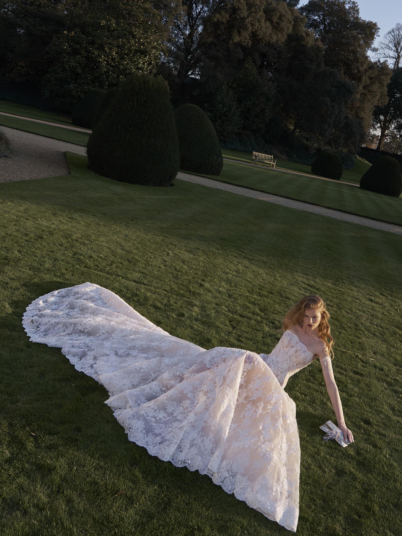 Woman in a white wedding dress with a long train lying on grass in a park.