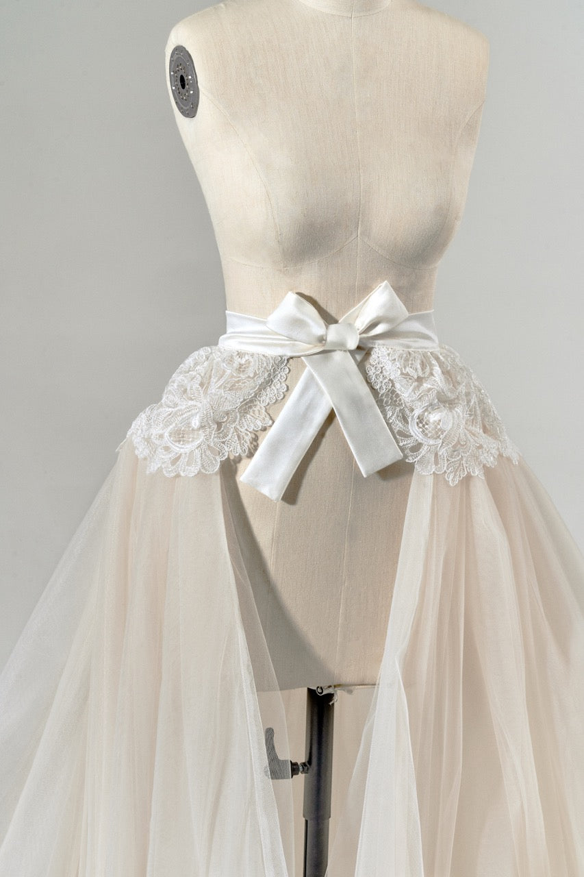 Mannequin wearing a beige tulle skirt with lace detailing and a white ribbon bow.