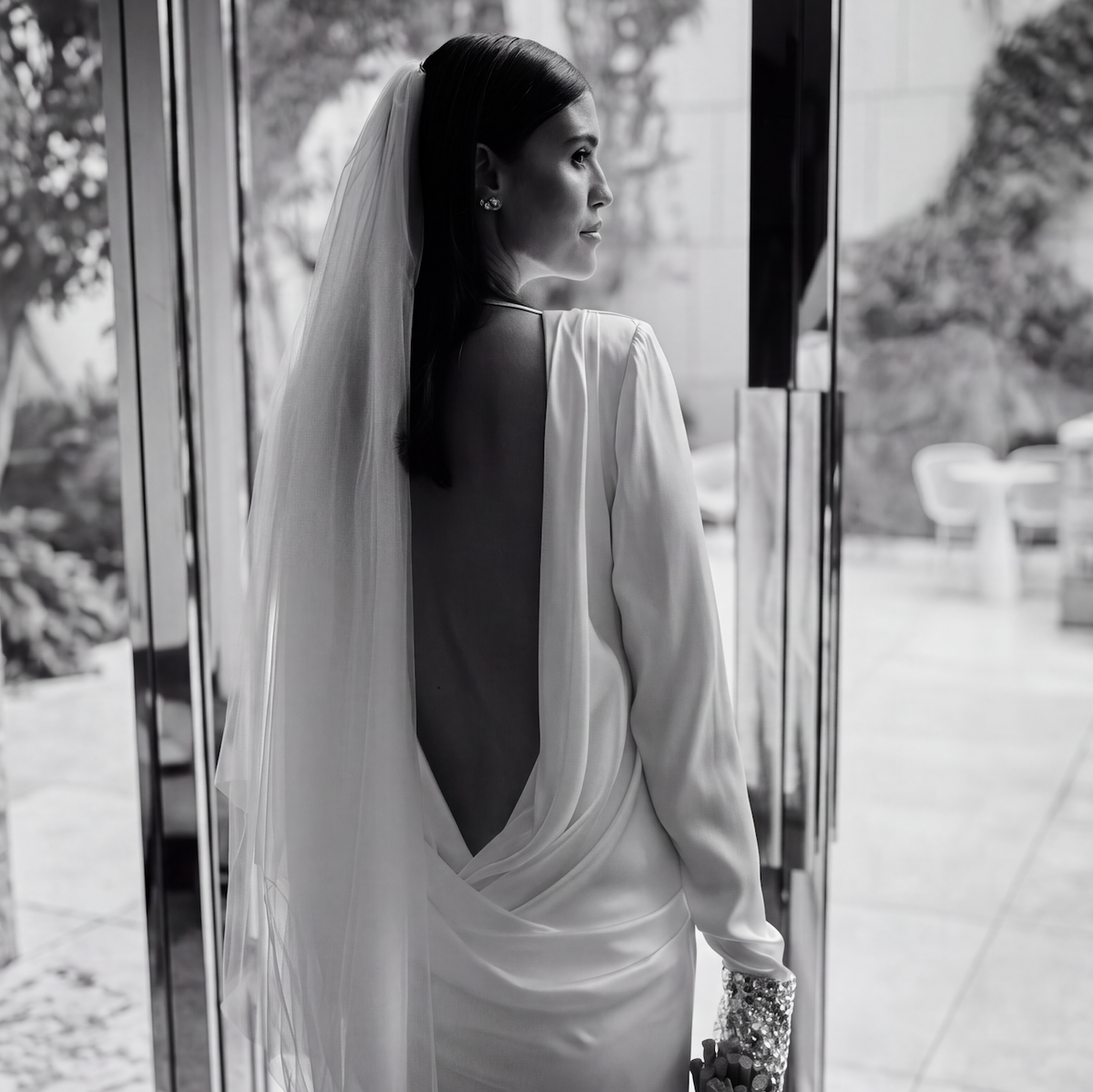 A black and white photograph of a woman wearing a long-sleeved backless wedding gown with a sheer veil, standing in front of a glass door and looking to the side.