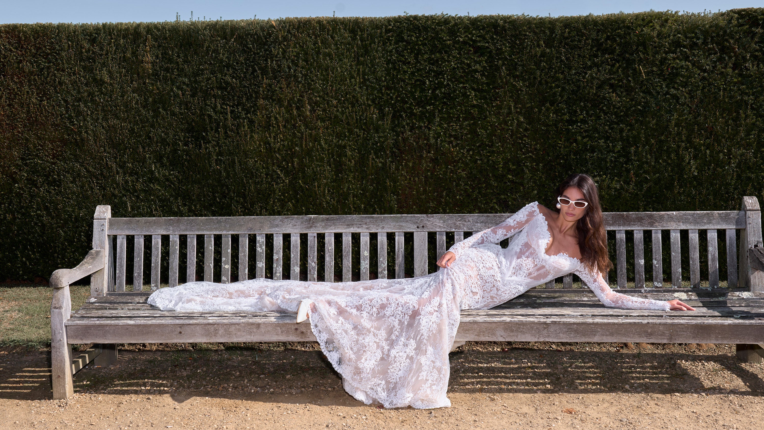 Woman in a white lace dress lying on a wooden bench against a hedge.