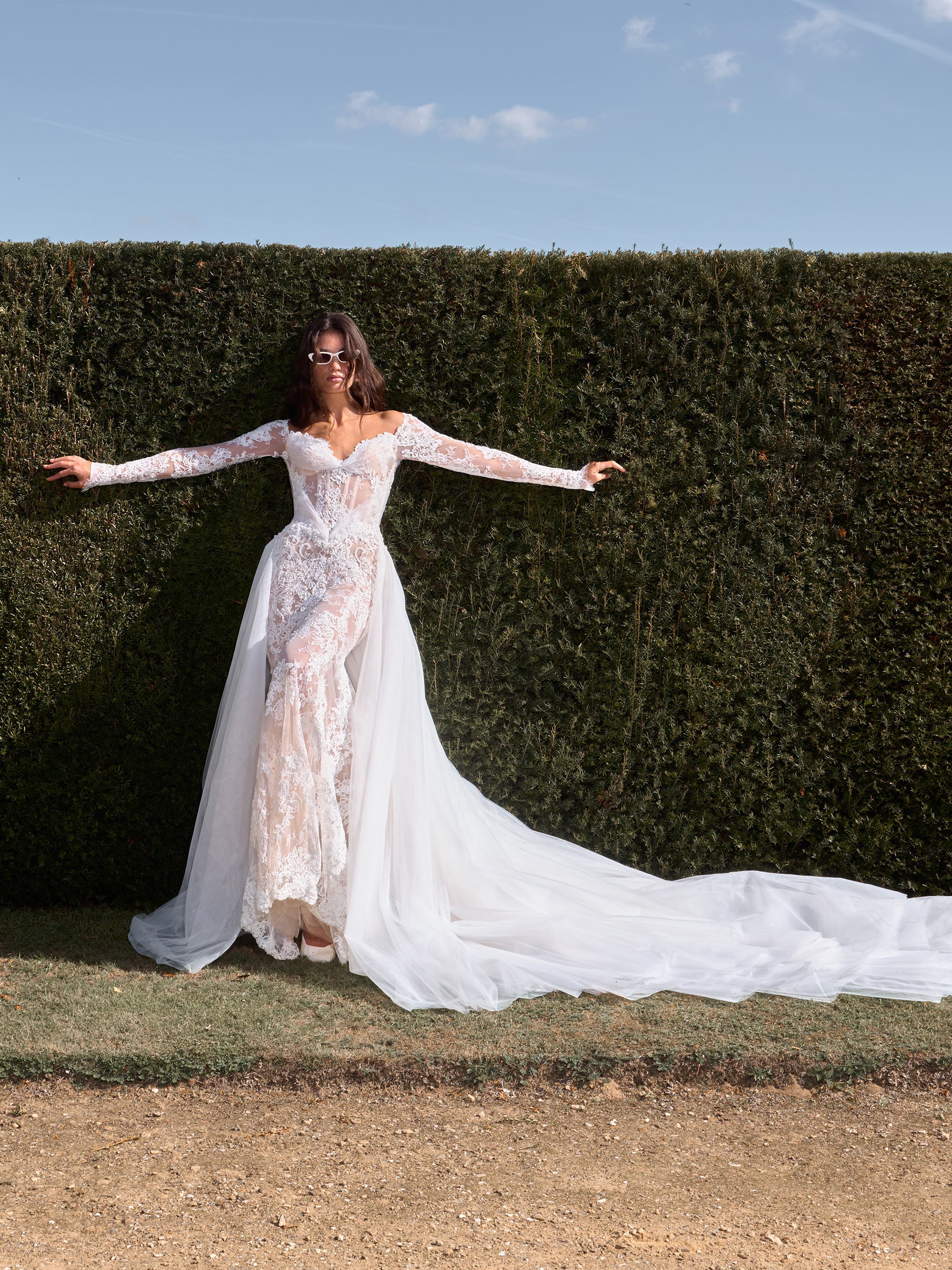 Woman in a white wedding dress standing in front of a hedge with a clear blue sky.