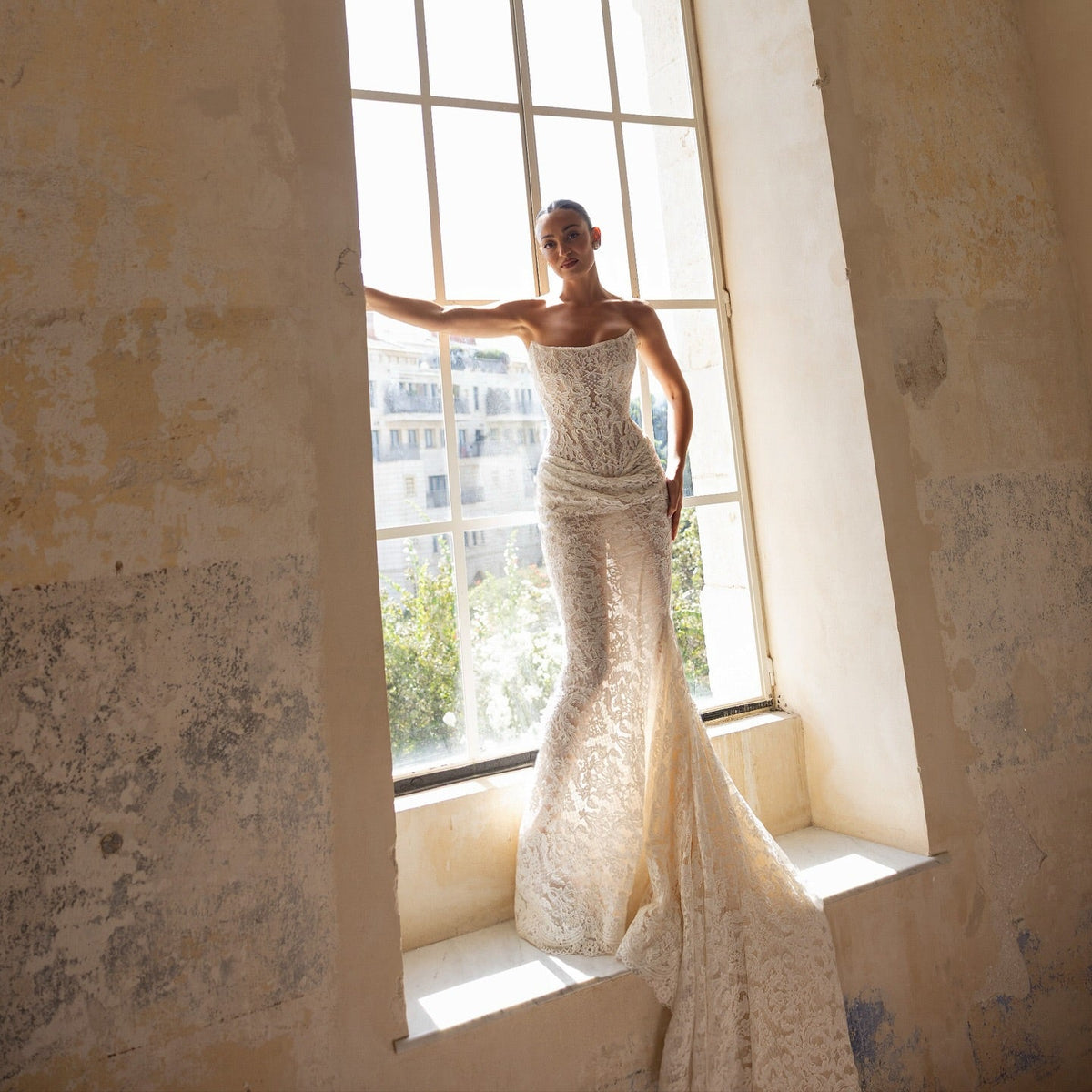 A woman wearing a strapless lace mermaid-style wedding gown stands by a large arched window in a sunlit room with textured beige walls.